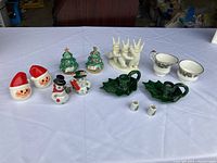 Christmas-themed salt and pepper shakers with Santa, snowmen and Christmas trees, green leaf candlestick holders, cream and sugar bowls, and white snow bunny decoration on white tablecloth