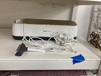 Photo of the Cricut Maker smart cutting machine with white power cable, USB cable, and scraper tool on a white shelf.