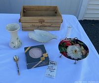Lot contents arranged on white tablecloth showing all items: wooden crate, Irish vase, Lenox bowl, terra cotta coasters, floral dish on stand, and vintage spoon.
