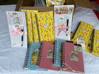 Photo showing multiple spiral notebooks and notepads with colorful, whimsical covers featuring cats, floral patterns, and floral tea themes, some with pens attached by pink ribbons.