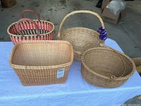 Four medium-sized wicker baskets displayed on a blue cloth, showing different shapes including rectangular, round with handle and flower, oval with damaged handle, and striped Shaker style.