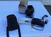 Photo showing the Pentax 8x24 UCF binoculars, box, carrying case, Pentax IQZoom 900 auto focus camera with zoom lens, lens cap, wrist strap, instruction manuals, and tags on a white surface.