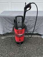 Front view of red and black Husky electric power washer, showing the whole unit with handle, power hose coiled around, and attached spray gun.