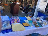 Wide shot of multiple quilting fabrics and materials spread out on a table in a garage or workshop. Shows a variety of fabric pieces and bundles in colors like blue, yellow, and floral patterns.