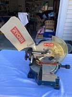 Front and side views of the RYOBI mitre compound saw showing the dust bag, blade guard, and adjustable handles on a white cloth surface outdoors.
