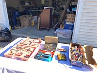 Wide photo of all items on white table outside garage including pink tool box with assorted tools, two staplers in packages, box of staples, and assorted screwdrivers.