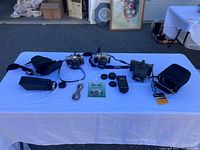 Wide view of the lot on a white table outdoors showing two film cameras, lenses, camera cases, flash, lens caps, and straps.