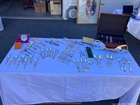 Wide view of vintage silverplated cutlery, Irish Donegal china spoons, vanity handheld mirrors, and silver cleaner displayed on a table outside in front of garage.