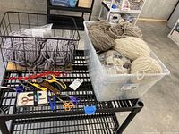 Full view of plastic bin containing bulky yarn skeins and various cutting tools on black metal shelf.