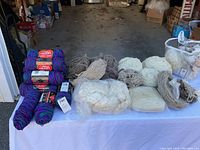 Wide shot of assortment of yarn and wool fibers on table including Red Heart Super Saver multicolor skeins and natural wool bundles.