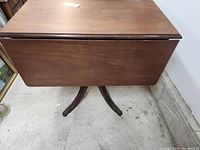 Side view of the wooden drop leaf table showing the closed drop leaf and the pedestal base with four ornately carved legs.