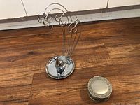 Full view of silver-tone peacock necklace display stand with circular ring case beside it on wood floor.