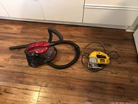 Photo showing a small black and red vacuum cleaner with hose and a yellow Dewalt jigsaw on the wooden floor.