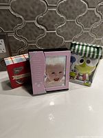 Photo showing pink baby photo frame featuring a baby, red box of metal puzzles, and boxed frog-themed toy