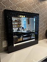 Black framed square mirror resting on a counter against a tiled backsplash, reflecting kitchen shelves and furniture.