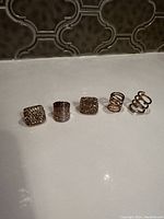 Photo showing five gold-tone costume rings arranged in a line on a white surface against a dark patterned background.