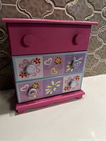 Front view of pink wooden jewellery box showing four drawers and hand painted decorative panels with hearts, flowers, and ladybugs.