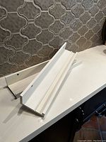 Two white IKEA floating shelves stacked on a white countertop with dark patterned tile background. Both shelves have mounting holes on the rear flange.