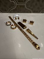 Photo of all items in set showing gold tone bracelet, tie clip, money clip, earrings, cufflinks, and rings arranged on white surface with tiled background.