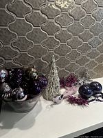 Various purple and silver Christmas bulbs arranged in a black container beside a white wire tree, silver wire tree, purple glitter floral picks, and a small white bird figurine.