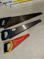 Photo shows three hand saws with varied sizes and handle colors laid on a white surface.