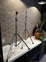 Two chrome-finished drum cymbal stands with tripod bases on a white surface against patterned tile wall.
