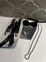 Black adjustable suspenders, black bow tie, patterned pocket square, and black beaded necklace laid out on white surface.