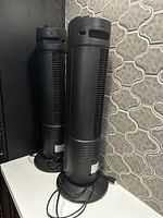 Two black oscillating standing tower fans shown side by side on the floor against a patterned wall. Fans have round bases, vented front panels, and are slim and tall.