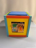 Front side of the baby activity box showing four square animal blocks (frog, tiger, others) inserted into the yellow face of the wooden box with blue and green trim.