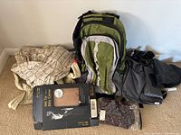 Green and gray baby carrying backpack, beige fabric baby wrap or carrier, black and gray diaper bag, and small dark accessory bag arranged on floor