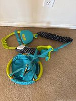 Top-down view of entire baby jumper showing blue fabric seat, green plastic frame, and black/white polka dot straps on carpeted floor.