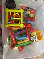 Plastic bin filled with various baby and toddler toys including a yellow and green activity cube with bead maze, ramps, balls, and other loose toy parts.