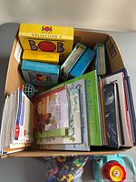 Top-down view of a cardboard box filled with assorted children's books including boxed Bob Books Collection 2, a green hardcover titled 'Minature', and several other books.