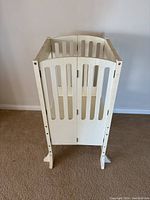 Front view of white wooden kitchen helper stool with tall side panels and slatted door-like front with two vertical cutouts to hold the child securely.