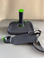 Overview of KneeGuard Kids 3 car seat foot rest showing height adjustment column and foot platform with green adjustment knobs, black textured surface, and attached strap with clip.