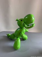 Right side view of green balloon-style dinosaur toy with visible head, teeth, legs, and tail on gray surface with neutral background