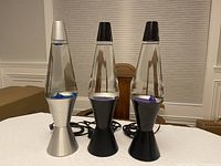 Three lava lamps displayed on a table: one silver with blue wax, two black with purple wax.