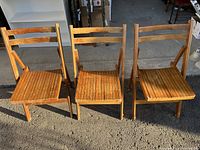 Three wooden folding chairs side by side on concrete floor with visible shadows.