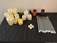 Top view of various battery operated candles arranged on a black surface, including flameless pillar candles, votive candles, tealights, Halloween-themed luminaries in pumpkin and skull shapes, and a set of long solar-powered candle-shaped LED lights with metal rods.
