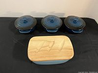 Butter dish with bamboo lid placed in front with three blue ceramic cocottes with lids behind it on a black surface.