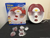 Foot spa device displayed next to its original packaging box, showing three sealed massage attachments in plastic and two reflexology plates inside the basin.