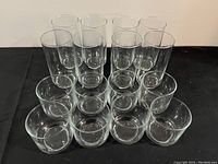 Photo showing 16 clear glass drinking glasses, arranged in rows with 8 taller glasses in the back and 8 shorter glasses in the front.