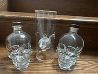 Photo shows two empty transparent glass skull-shaped decanters with black screw caps placed on wooden steps with a small glass oil lamp inside a clear cylindrical container between them.