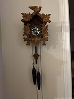 Front view of the wooden German cuckoo clock mounted on a wall showing carved leaves, a bird, black clock face with white roman numerals and two pinecone weights hanging.