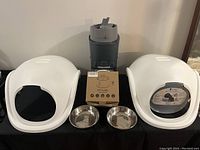 Lot displayed on a table: two white litter locker hoods, two stainless steel bowls, sealed pet fountain in box, and automatic food feeder