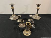 Full view of 6 metal candlestick holders, 2 tall and 4 shorter, arranged on black surface.