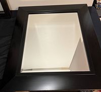 Black framed rectangular wall mirror with wide frame shown lying flat on a black fabric surface.