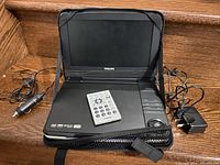 Philips portable DVD player open to show screen and control panel, with remote on top and two adapters on sides, all placed on wood surface.