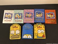 Top down view of eight DVD box sets of The Simpsons seasons 1 through 8, five in rectangular cardboard sleeves and three in shaped metal cases of Homer, Marge, and Bart Simpson heads.