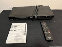 Black Toshiba Blu-ray player with power cord, remote, and user manual laid out on black surface.